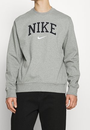 Mann trägt grauen Nike-Sweatshirt mit schwarzen Buchstaben und weißem Swoosh-Logo, kombiniert mit schwarzen Hosen, vor einfachem Hintergrund.