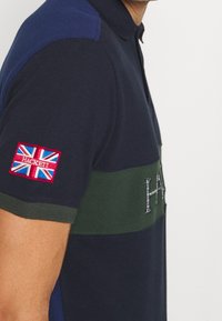 Marinblå polo-shirt med grön horisontell rand; har ett rött och blått broderat märke med den brittiska flaggan och "Hackett"-varumärket.