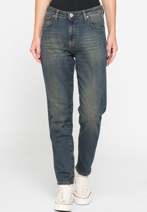 Adulto che indossa jeans blu slavati slim fit e sneakers bianche alte, in piedi su uno sfondo bianco uniforme.