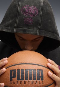 Czarna bluza z kapturem z fioletowym haftowanym logo, trzymająca pomarańczową teksturowaną piłkę do koszykówki z wyraźnym napisem marki. Zbliżenie na dłonie i twarz.