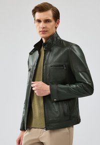 Giacca in pelle verde scuro con colletto alto, apertura con zip e numerose tasche. Texture liscia con dettagli di cuciture accentuate.