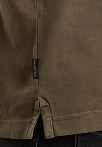 Olivengrøn bomulds-T-shirt med sideslidser, der har et lille sort mærke med teksten "Dame Legend" nær kanten. Glat tekstur, tætsiddende design.