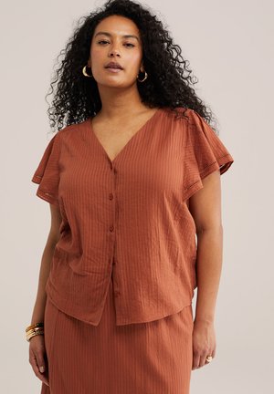 Femme aux cheveux noirs bouclés portant une blouse boutonnée couleur rouille et une jupe assortie, avec des boucles d'oreilles anneaux dorés et des bracelets sur un fond beige.