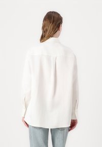 Camicia bianca a maniche lunghe con colletto, taglio dritto e discreto pieghettato sul retro. Il tessuto appare leggero e liscio. Abbinata a jeans azzurri chiari.
