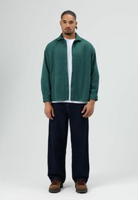 Dunkelgrünes Hemd aus strukturiertem Stoff, getragen über einem weißen T-Shirt. Kombiniert mit locker sitzenden dunklen Jeans und braunem Schuhwerk.