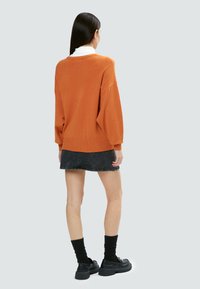 Lange, orangefarbene Sweater mit lockerer Passform, gerippten Bündchen und Saum, kombiniert mit einem dunklen Jeansrock und klobigen schwarzen Schuhen.