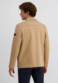 Beige katoenen blendjas met een gestructureerde kraag, lange mouwen en een logo patch op de linkermouw. Kenmerkt zich door strakke lijnen en een minimalistisch ontwerp.