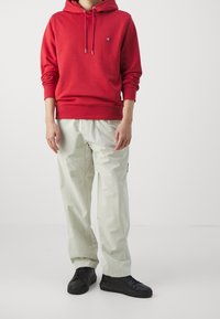 Sudadera con capucha roja y cordones, logo en el pecho, combinada con pantalones de utility gris claro y zapatillas negras.