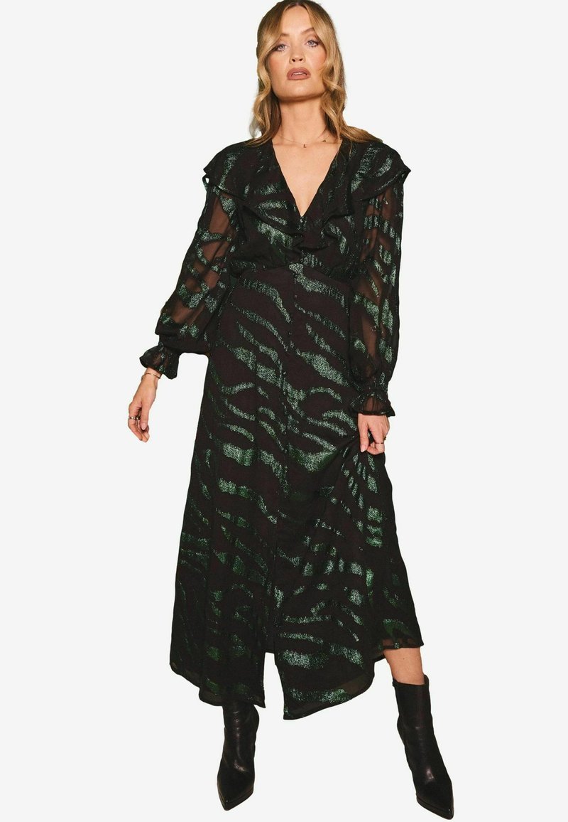 Rochie maxi din material negru transparent cu un model de dungi de tigru verzi, cu un decolteu volanat, mâneci lungi și o siluetă fluidă. Stilizată cu cizme negre.