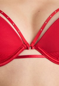 Raudona liemenėlė su tinklelio audiniu, trikampiais puodeliais ir dekoratyviais dirželiais su mažais metaliniais akcentais. Kombinuojami tekstūruoti ir lygaus paviršiaus audiniai.