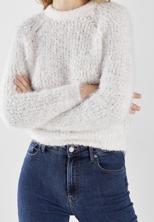 Personne portant un pull blanc duveteux à manches longues et un jean bleu taille haute, les bras croisés.