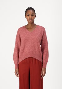 Maglione lavorato a maglia rosa con scollo a V, spalle scese e orlo a coste. Indossato sopra pantaloni rossi a gamba larga, con una leggera texture e calore.