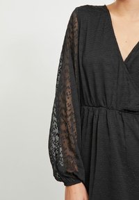 Svart klänning med en texturerad, enfärgad bodice och genomskinliga puffärmar, som har ett subtilt mönster. V-ringad design med elastisk midja.