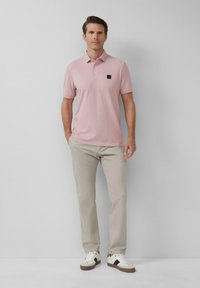 Polo shirt rose clair avec un patch logo noir, associé à un pantalon beige et des baskets blanches avec des accents bruns et des lacets plats.