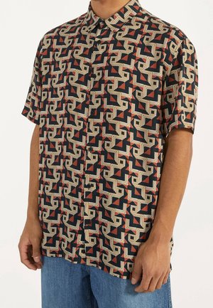 Homme portant une chemise à manches courtes à boutons avec un motif géométrique en zigzag noir, beige et rouille, associée à un jean bleu.