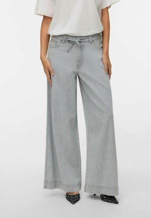 Kvinne iført lysegrå vide jeans med knyttet midje og svarte spisse hæler med hvite polkadotter, delvis synlig overkropp.