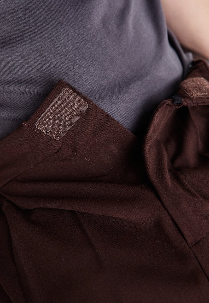 Pantalons en tissu marron avec une texture lisse, dotés d'une patte en Velcro et d'une fermeture à bouton à la taille, associés à une chemise grise.