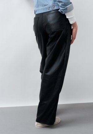 Άτομο που φοράει μαύρο δερμάτινο παντελόνι με φαρδιά μπατζάκια, μπλε ριγέ σακάκι και λευκά παπούτσια, στέκεται μπροστά σε απλό ανοιχτό φόντο.