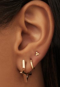 Boucles d'oreilles en ton doré sur une oreille : l'une présente un design géométrique en forme d'éclair ; l'autre est une petite créole avec trois accents de perles.
