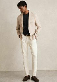 Sacou bej deschis peste un tricou negru, pantaloni albi și mocasini maro. Materialul este neted, iar croiala este ajustată.