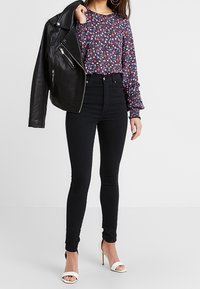 Femme portant un jean skinny taille haute noir, un chemisier à manches longues à fleurs, des sandales blanches à talons, et tenant une veste en cuir noire sur une épaule.