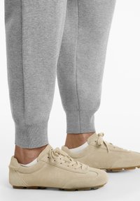 Beige velúr cipők kerek orral és fűzős elülső résszel, szürke, tapered szabású melegítőnadrággal párosítva, amely bordázott mandzsettákkal és sima textúrával rendelkezik.