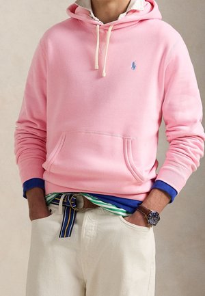 Person trägt einen pinken Hoodie über mehreren Schichten von Hemden, weiße Hose mit gestreiftem Gürtel und eine Armbanduhr, Hände in den Taschen.