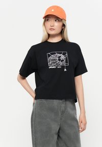 Black cotton t-shirt with a white spider web graphic and text. Paired with grey patterned pants and an orange cap.