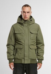 Chaqueta aislante verde oliva con capucha, cuatro bolsillos frontales y puños de canalé. Presenta bolsillos laterales con cremallera y una textura suave.