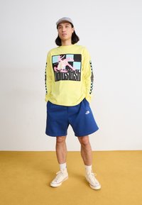 Φωτεινό κίτρινο μακρυμάνικο πουκάμισο με γραφικό σχέδιο, μπλε σορτς και κρεμ αθλητικά παπούτσια. Γκρίζο καπέλο καρό, casual στυλ, απλό φόντο.