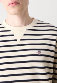 Stripet langermet skjorte i kremfarge med marineblå horisontale striper. Ribbet tekstur, rund hals og en liten brodert logo på brystet.