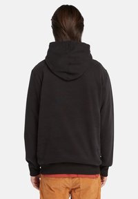 Persona con capelli castani di lunghezza media, che indossa un hoodie nero e pantaloni arancioni, vista da dietro su uno sfondo neutro.