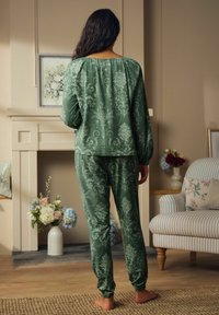Zaļš rakstains pidžamu komplekts ar garām piedurknēm un elastīgām aprocēm, ar ziedu dizainu, apvienots ar atbilstošam biksēm, skatīts no aizmugures.