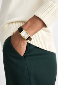 Montre-bracelet à cadran carré avec un boîtier doré, un cadran blanc, un bracelet en cuir noir et des finitions en argent, portée avec un pull crème et un pantalon vert.