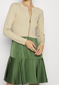 Cardigan beige in maglia con zip frontale e texture a motivo, abbinato a un vestito in satin verde a strati con orlo svasato.