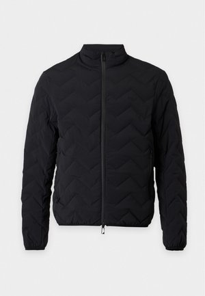 Homme debout portant une veste matelassée noire par-dessus un polo noir, un jean foncé et des baskets noires sur un fond clair uni.