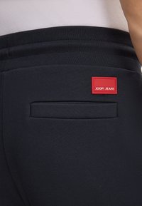 Schwarze Jogginghose mit glatter Textur, elastischem Bund und einer Gesäßtasche. Ein rotes Etikett mit "JOOP! JEANS" ist sichtbar.