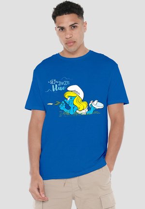 Man wearing blue T-shirt with cartoon character lying on grass and text "the sky is always blue" on front.