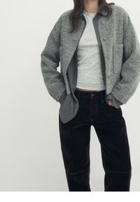 Veste en laine grise avec grandes poches, fermeture à boutons, superposée sur un haut gris clair et un pantalon en velours côtelé marron. Texture douce et coupe décontractée.