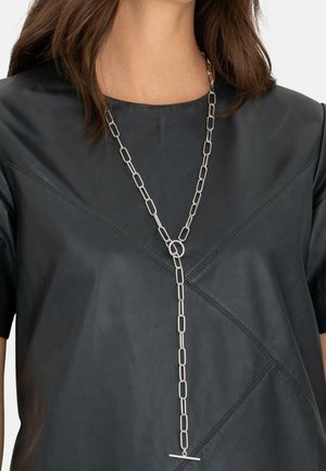Femme portant un collier argenté allongé à maillons rectangulaires sur un haut en cuir noir avec des coutures diagonales.