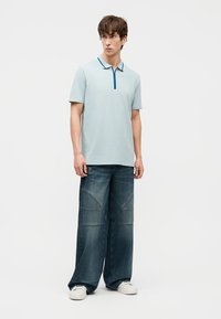 Jeune homme portant un polo bleu clair à manches courtes, un jean large foncé avec des détails cousus, et des baskets blanches, debout et regardant sur le côté.