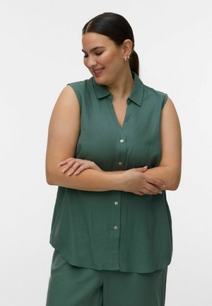 Kobieta o ciemnych włosach związanych w niskiego kucyka, ubrana w zieloną koszulę bez rękawów na guziki i dopasowane spodnie, z założonymi skrzyżowanymi ramionami, delikatnie się uśmiechająca.