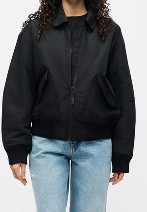 Schwarze Jacke mit Reißverschluss und zwei vorne schräg angebrachten Klappentaschen, getragen über einem schwarzen Hemd und kombiniert mit hellblauen Jeans.