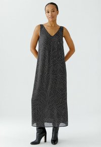 Robe maxi noire sans manches avec un décolleté en V, ornée d'un motif ondulé blanc et d'un tissu léger et fluide. Associée à des bottes à talons noires.