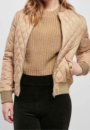 Femme portant une veste matelassée beige sur un pull en maille marron clair et un pantalon noir, une main dans la poche de la veste.