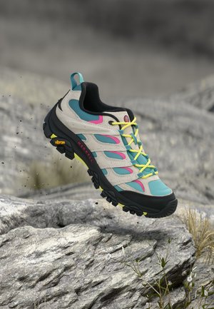 Lichtblauwe en beige wandelschoen met felgele veters en zwarte zool zweeft boven een rotsachtige ondergrond met kleine planten.