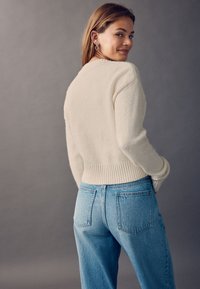 Cremefarbener, strukturierter Pullover mit einem gerippten Saum, kombiniert mit hellblauen, hoch taillierten Jeans mit zwei Gesäßtaschen. Einfaches Design.