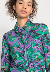 Camisa colorida de botões com uma base verde e padrões abstratos em roxo, laranja e preto, mangas longas e um design de colarinho clássico.