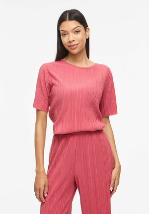 Femme aux longs cheveux foncés portant un haut plissé rose à manches courtes assorti et un pantalon, debout devant un fond blanc.