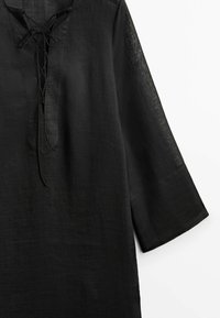 Tunique noire à manches longues avec un col en V orné de liens croisés à lacets et un tissu ample légèrement texturé.